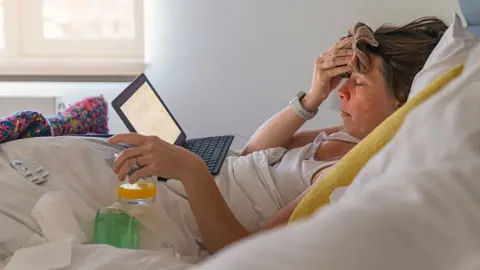 Getty Images A women in bed . she is unwell and has a flannel on her head and drinking a glass of orange