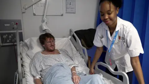 A nurse stands beside a hospital bed while a patient sits up under a blanket.