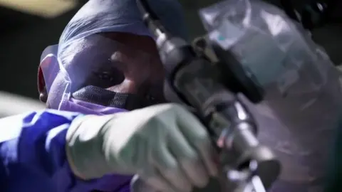 Sivakumar Shankar is seen wearing a surgical mask, cap and gloves looks through a piece of medical equipment while holding it with one hand.