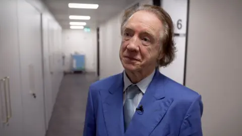 BBC An older man wearing a blue suit with shirt and light blue tie. He has brushed back brown hair and looks off to his right side