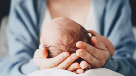 Getty Images A mother resting a baby's head in the palm of her hands, on her lap, so that we can see only the back of the infant's head. The mother is wearing a blue cardigan.