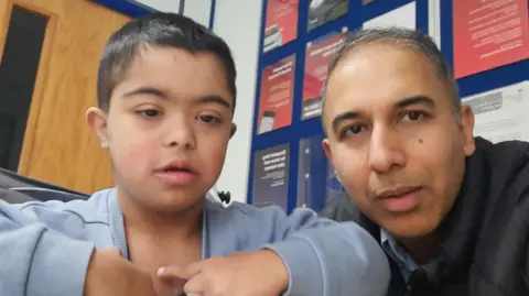 Family handout The picture shows two people sitting indoors. One person is wearing a light blue zip-up top and is pointing towards their chest with both hands. The other person is dressed in a dark padded jacket. Behind them, there is a blue noticeboard filled with various posters and information sheets, mostly in red and white colours. To the left, there is a wooden door.