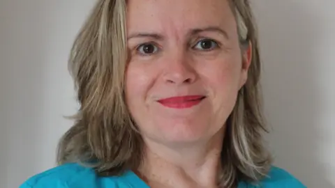 Dr Niamh Lynch A woman with long grey and blonde hair is smiling to the camera. She is wearing light blue hospital scrubs with her name across the right breast pocket. 