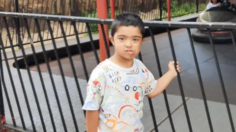 Family handout A young boy with dark hair and a white t-shirt is standing in front of dodgems at the fair, he is holding onto the railing and looking at the camera.