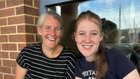 Maria Horton A mum and teenage daughter smiling, sitting in front of a redbrick building, with a window in the background. The mother wears a black and white striped t-shirt and the daughter wears a navy blue t-shirt with white logo. 