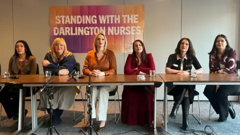 Six women sit at a long table with microphones set up in front of them. Behind them is a banner with the words "standing with the Darlington nurses" written on it