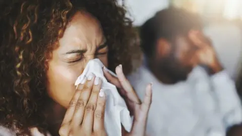 Getty Images A Woman with curly hair is sneezing into a tissue. There's a man in the blurred background behind the woman who has his head in his hands. 