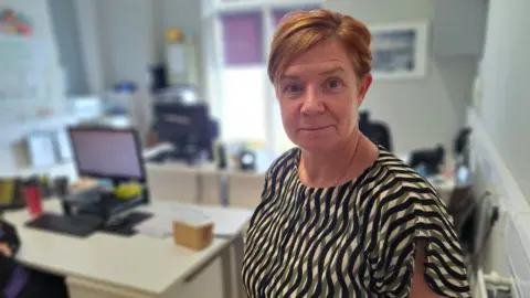 Cheryl Williams is stood in an office with desks and computers behind her. She has short auburn hair and is wearing a black, green and cream striped top and is looking directly at the camera. 