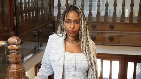 BBC Kiki Marriott is stood inside the Parkland Place Rehabilitation Centre at the top of some wooden stairs. She has blonde braids and is wearing a white patterned suit. Kiki looks directly at the camera.