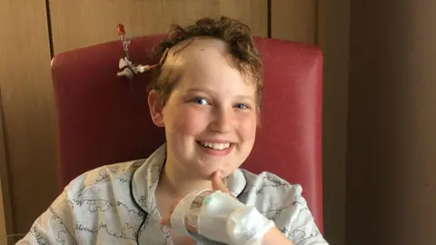 Louise Dempster A young girl who is smiling at the camera. She is sitting in a red chair and is wearing pyjamas. Her left hand is wrapped in a bandage and appears to be covering a canula. Her head has been partly shaved and some kind of medical contraption is sticking out of the right side of her head. 