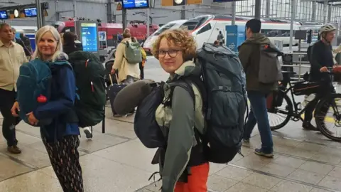 Louise Dempster A teenage girl standing in a train station with a large rucksack on her back, and a smaller one on her front. She is wearing dark-rimmed glasses and has her head turned to smile at the camera. 