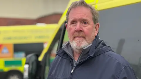 A photo of a man with dark grey hair and a beard, wearing a black rain coat, stands in front of an ambulance.