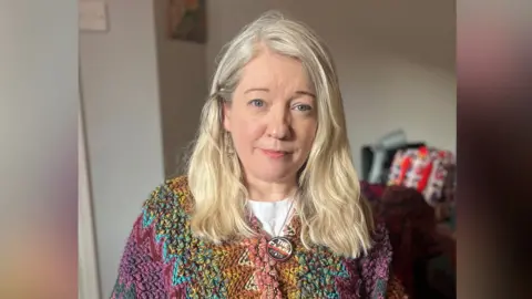 Marie Mullan A woman, pictured from the torso up, with long blonde hair. She has a multicoloured woven top on and a 'The Clash' necklace. There is a blur on either side of the image. 