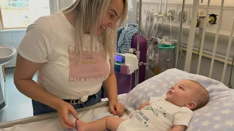 Samantha Williams Samantha Williams and her baby son Lucian Neale are in a hospital room. Lucian is lying down on the bed, with his body and head resting on a large pillow. He's looking up at his mum Samantha, who has blonde hair and is smiling back at him.