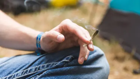 Getty Images A close-up stock photo that shows the knee and hand of a man wearing jeans sitting cross legged. He's holding what appears to be a half-smoked cannabis joint.
