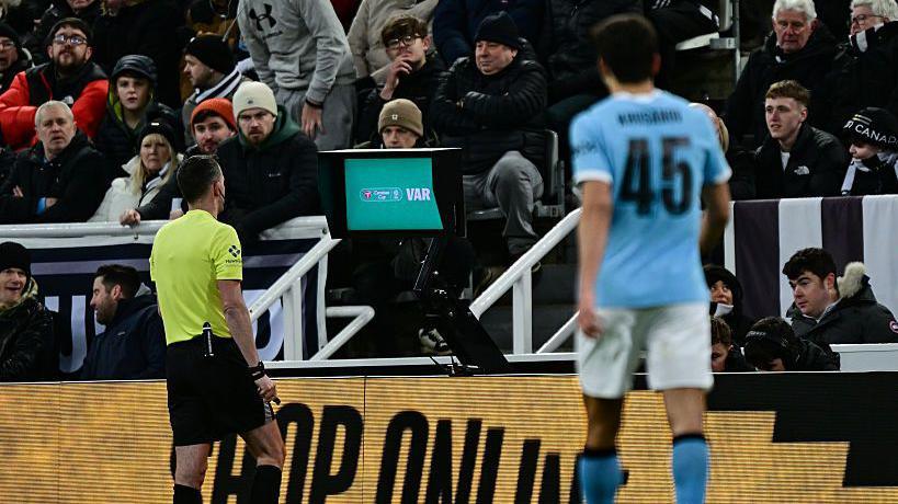 Referee Chris Kavanagh watches the pitchside VAR monitor, with crowd in the background