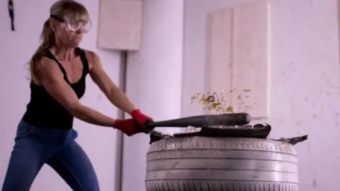 Getty Images Picture of a woman in a rage room smashing tyres with baseball bats. She is wearing a visor, black vest top and jeans.