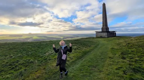 Cathy Read Woman holds her hands up with peace signs and she is on top of a hill with a large obelisk-style structure behind her.