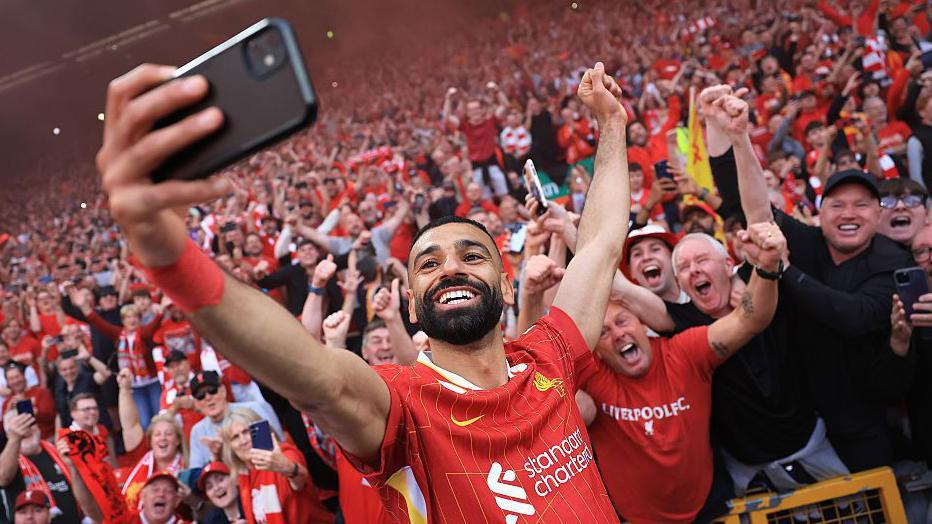 A grinning Mohamed Salah, in red club kit, takes a selfie with a mobile phone in front of excited Liverpool supporters