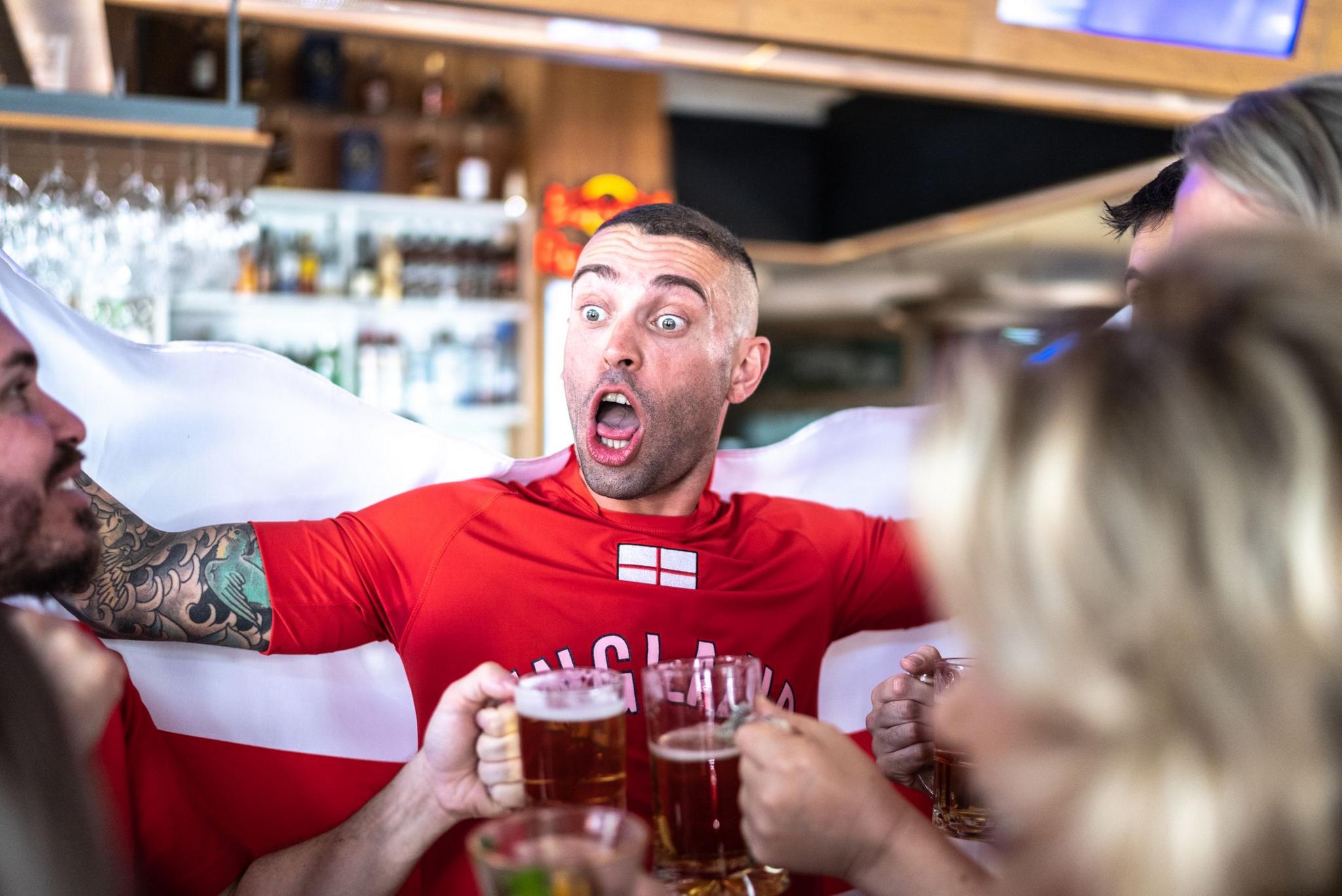 A young man, wearing an England T-shirt, is celebrating in the pub with friends. He is lifting an England flag behind him, worn like a cape. His friends are chinking pints of beer. 