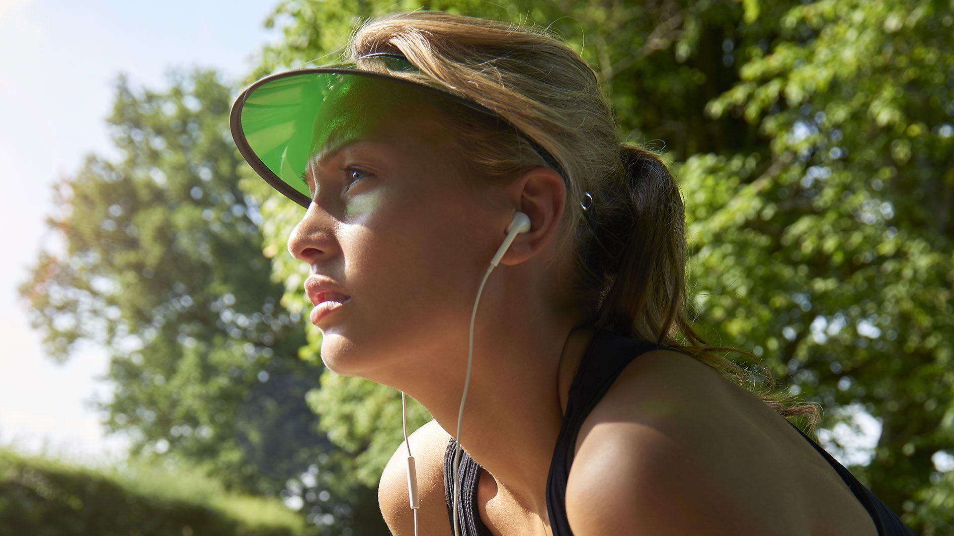 A young woman with a green visor and white earbuds squints into the distance in the countryside