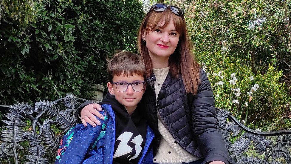 A portrait photo of Dima and Elena staring at the camera while sitting on a park bench. He is waring a blue jacket and has glasses. She is wearing a black jacket and white jumper and has sunglasses on her head.