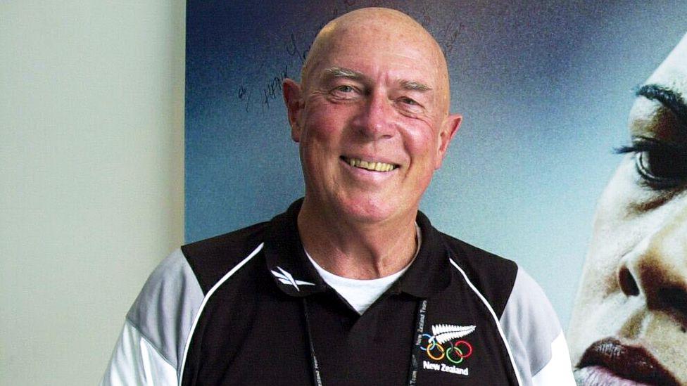 Les Mills, a man with bushy greying eyebrows and a bald head who is smiling straight ahead - he is wearing a black, white and grey sports top which has the Olympic rings, a silver fern logo and the words 'New Zealand' on the right-hand-side - he is standing in front of a poster, on which part of a woman's face can be seen 