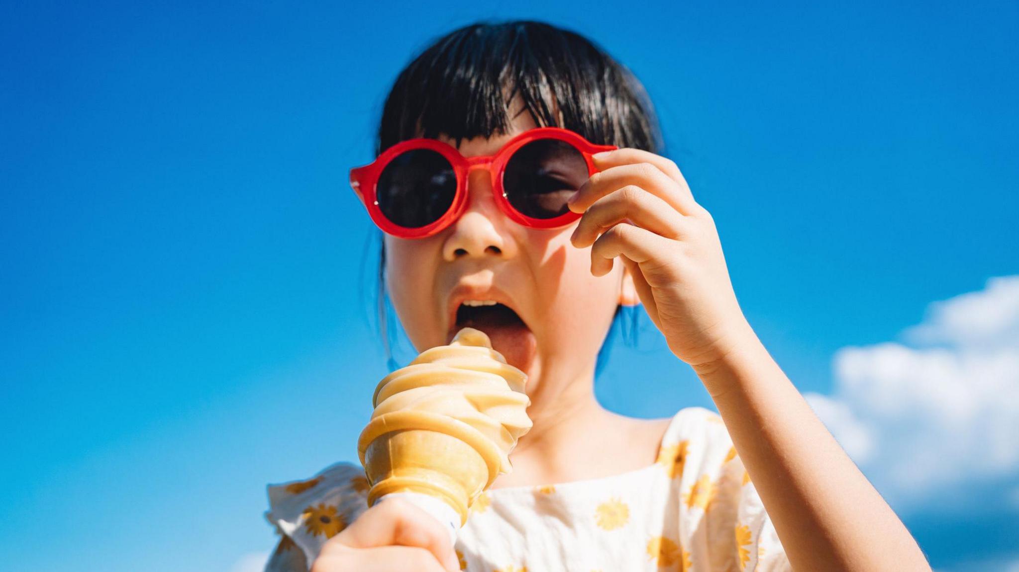 Young girl with dark hair wearing red sunglasses and eating ice-cream cone outdoors on a sunny summer day against blue sky