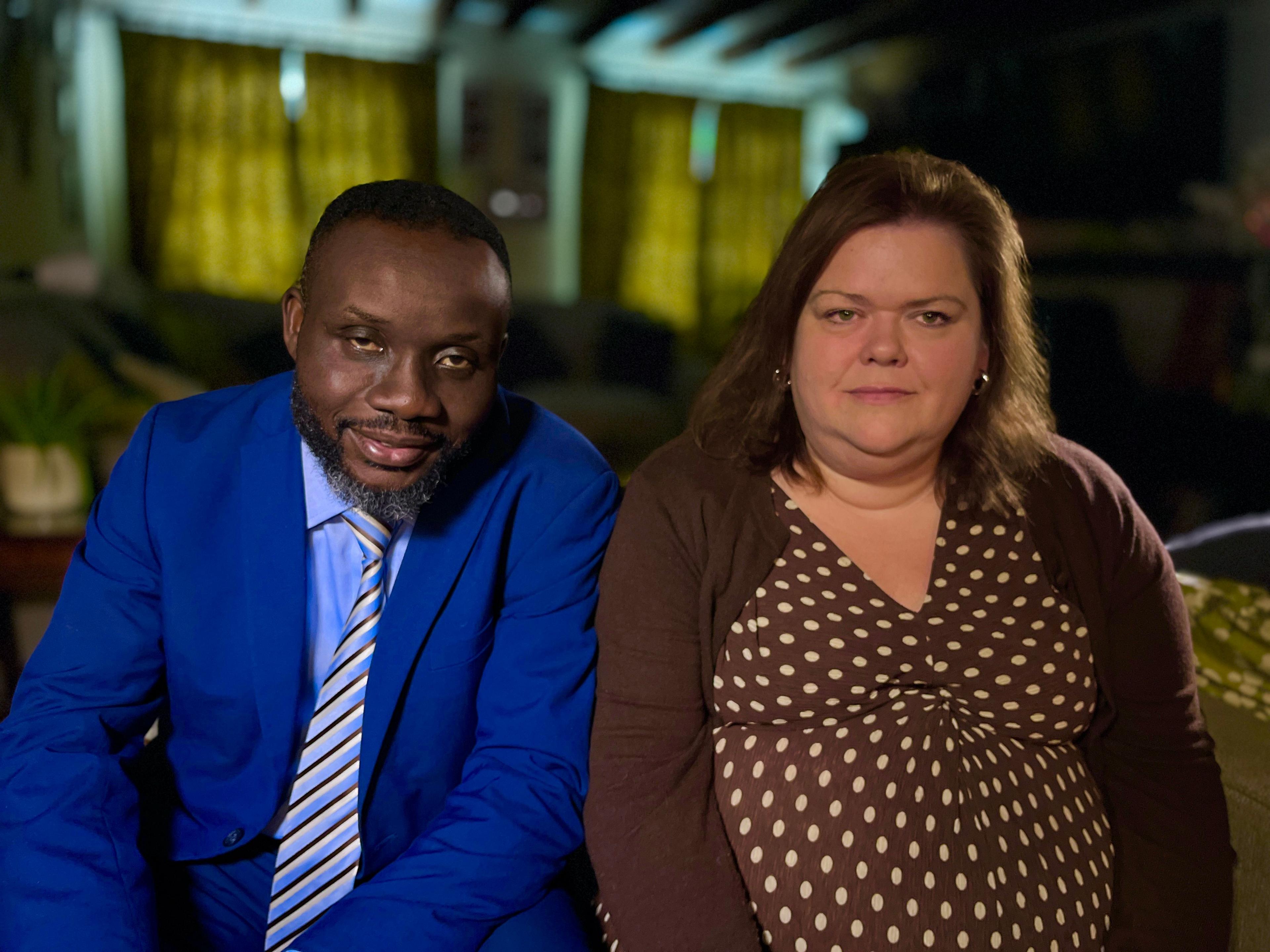 Caroline with her husband Lionel, both look at the camera. Lionel is on the left, wearing a royal blue suit with a blue, yellow and black striped tie. Caroline is wearing a brown spotted dress with a chocolate brown cardigan.