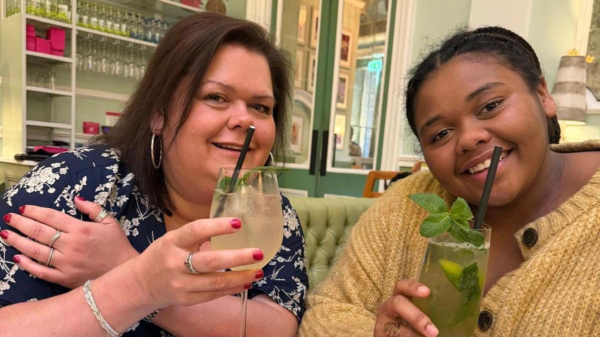 Caroline with her daughter Francesca. They are both holding Mojito cocktails. One is non-alcoholic. Caroline has a navy blue flowery dress and silver hooped earrings and Francesca has a fluffy yellow cardigan. They are in a bar.