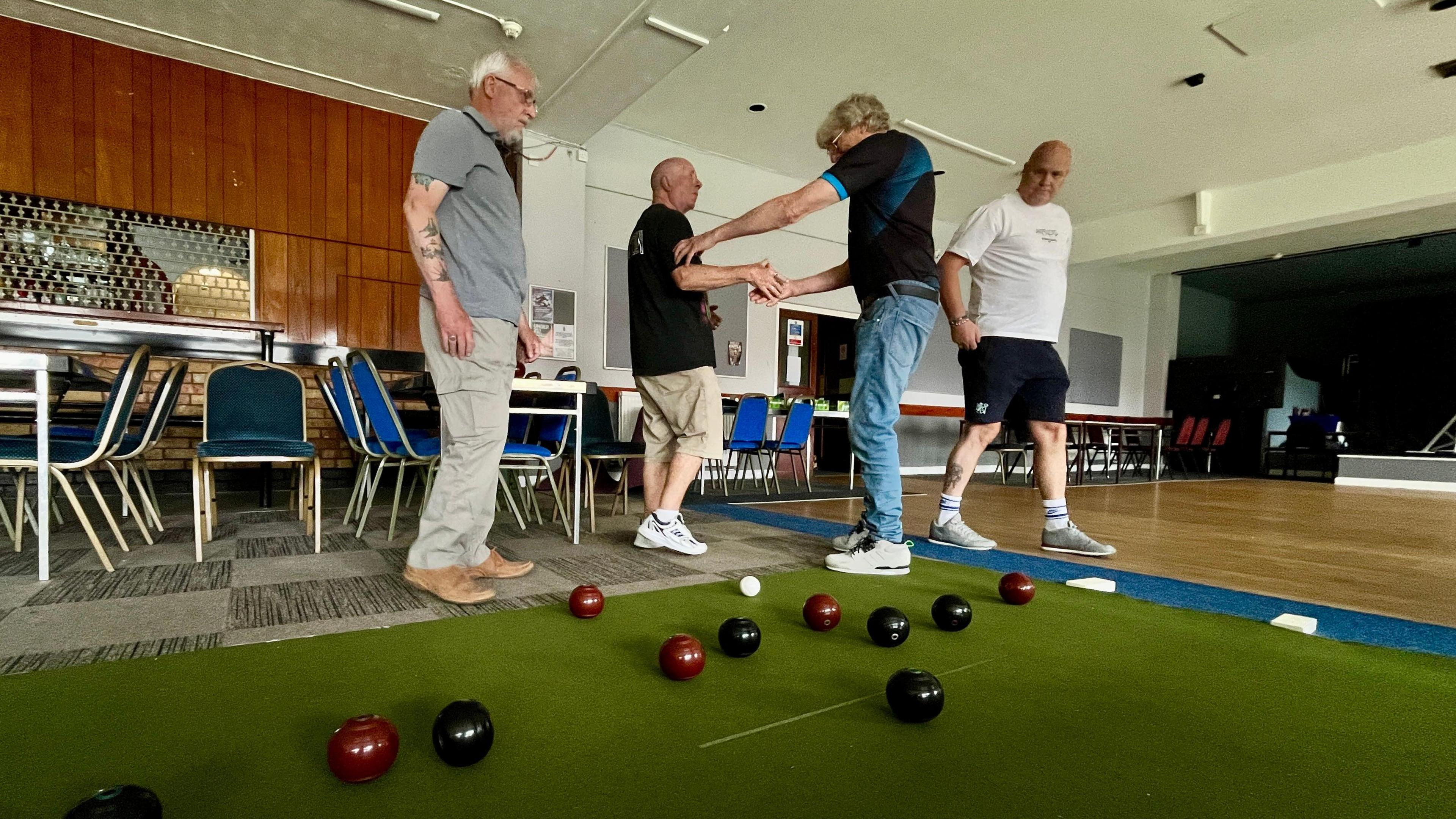 A group of four men playing short mat bowls at the Montagu working man's club in Huntingdon. Shane is on the right wearing a white t-shirt and black shorts. Two men are shaking hands and congratulating themselves in the middle. A man on the right is looking at the bowls on the floor, as is Shane. There are velvet covered function chairs in the background, and the bar shutters are down. There is a small stage to the right.