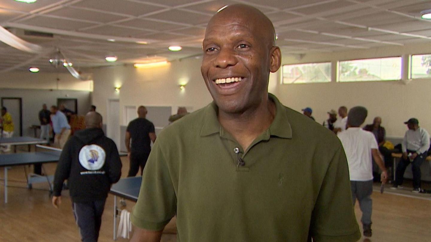 Chidi Umeano standing in a room, wearing a green top, with men playing table tennis behind him. There are windows in the room. He is smiling and looking off to the camera. 