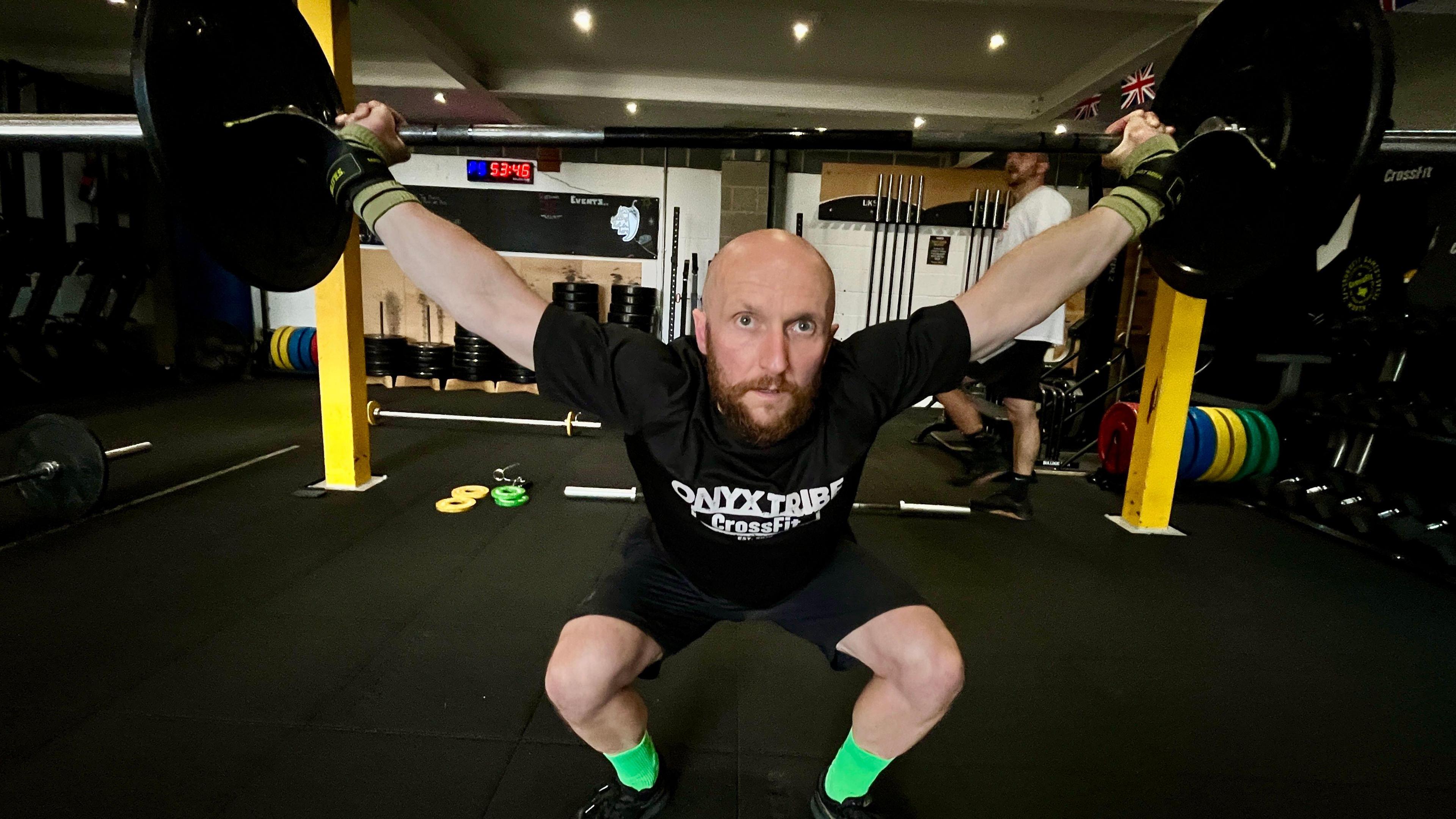 Lukas Koziel picks up weights on a bar above his head in a snatch. He has a brown beard and bright green socks. There are lots of weights in the background. He has grips on his hands and his arms are spread above him in a v shape. He has a black t shirt and shorts on.