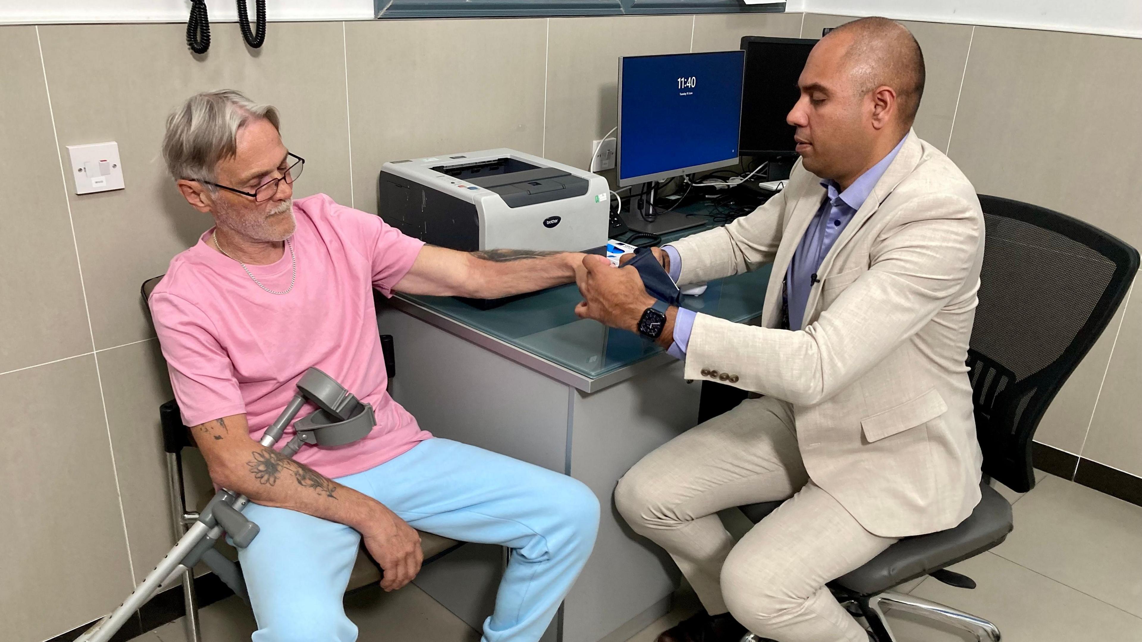 Dr Neil Modha examines a male patient who is in his 60's. Dr Modha has a beige suit on. He is putting a blood pressure cuff on the patient while sitting to the side of his desk with a computer and printer on. The patient has a pink t-shirt and pale blue jogging bottoms. He has glasses on and crutches across his lap.