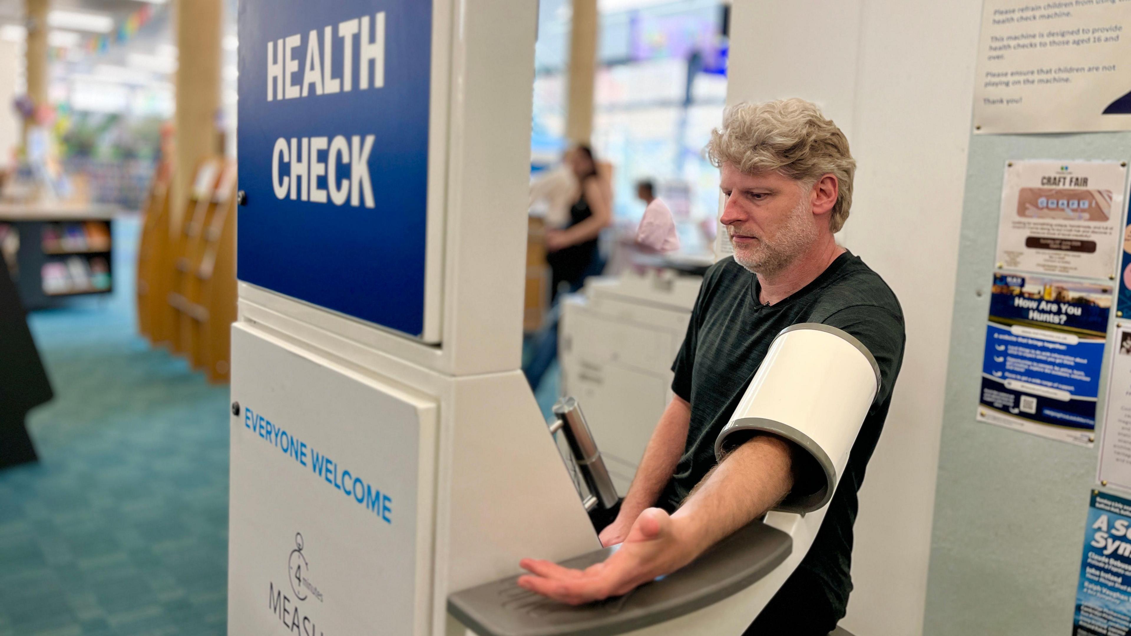 Michael Hadley has his arm through a blood pressure monitor cuff. It is attached to the machine and is a tube which has the monitor cuff inside it. It inflates as he rests his arm on a plastic ledge. The machine is labelled 'free health check', You can see some library shelves in the background