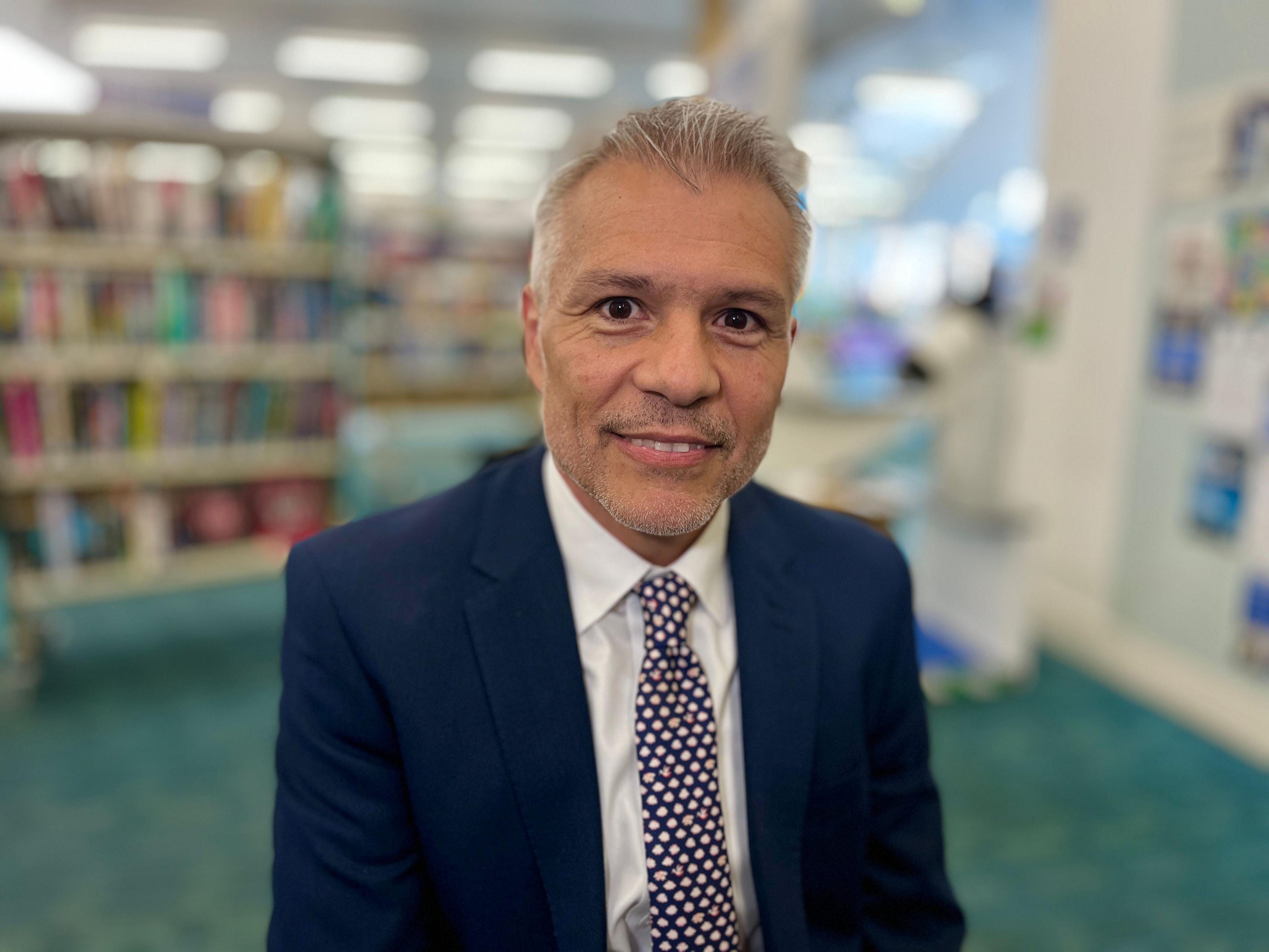 Councillor Louis Navarro looks at the camera wearing a navy suit with a white shirt. He has a spotty tie on and is smiling. He is standing in the library in Huntingdon.