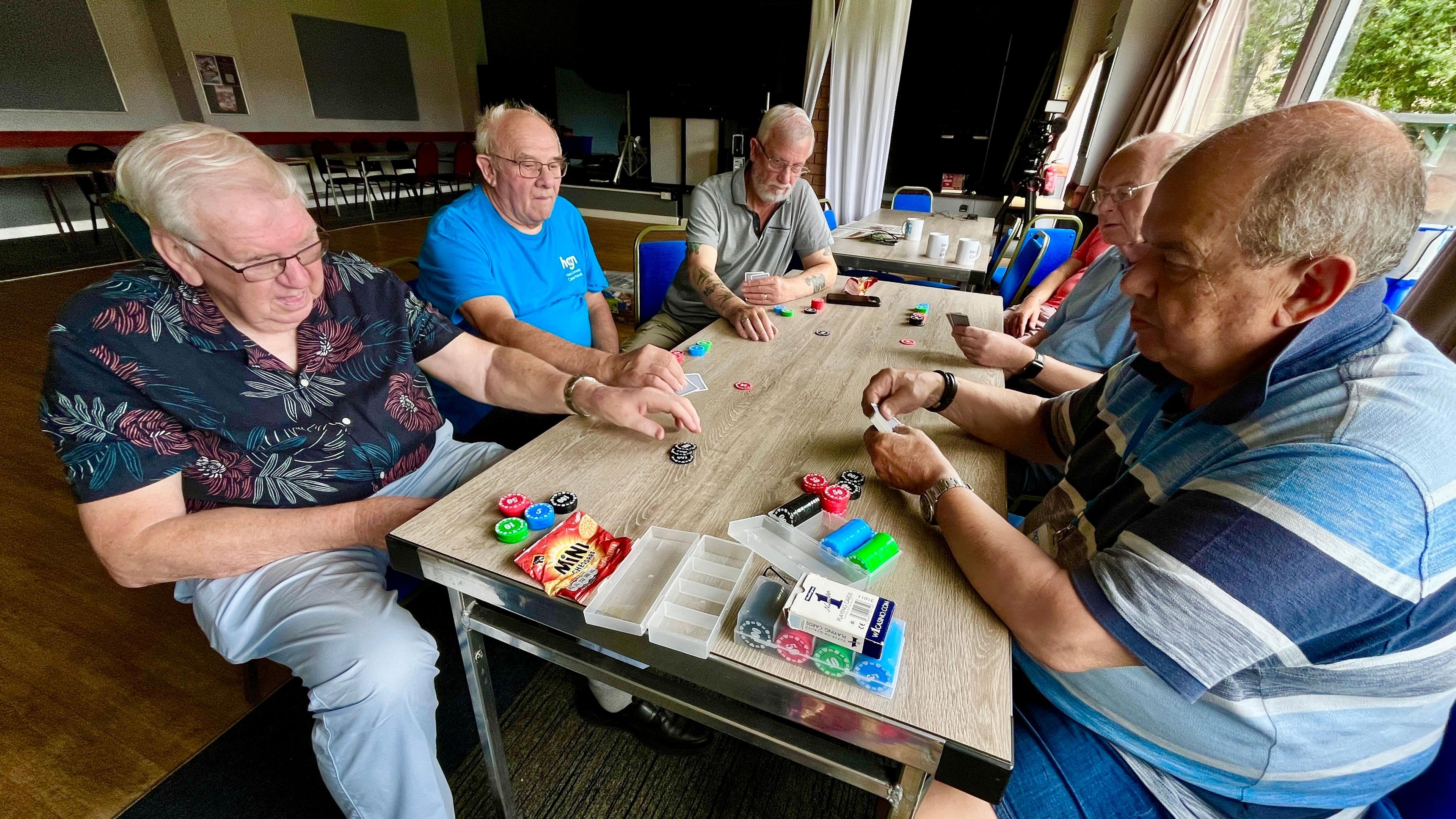 A group of six men sit round a table at the working man's club playing cards. They have counters that they are using to place bets. They are largely retired and are wearing t-shirts or light summer shirts
