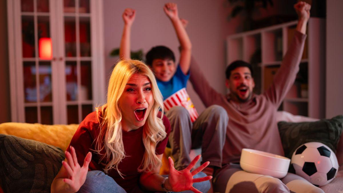 A blonde woman watches football on sofa next to a child and a man. There's a football next to them. The man has a bowl on his lap. A door and a bookshelf are visible in the background