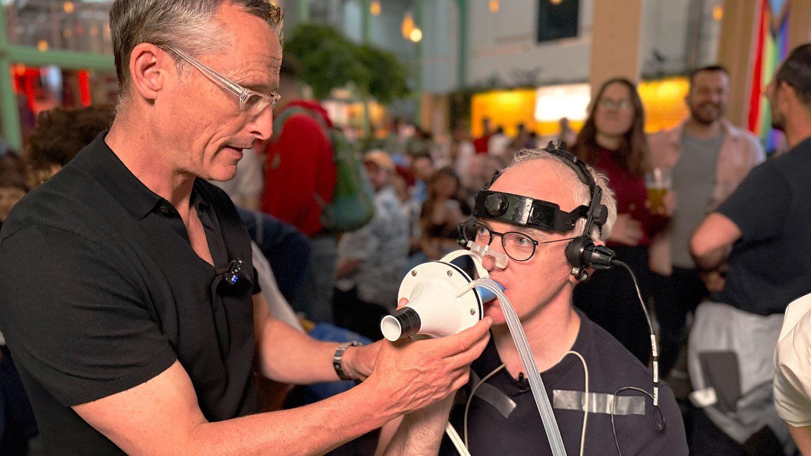 Prof Damian Bailey holds a white breathing mask connected to clear tubing up to James' face, who is seated and wearing a head-mounted device with wires and sensors attached. James has additional medical-style electrodes taped to the chest and arm