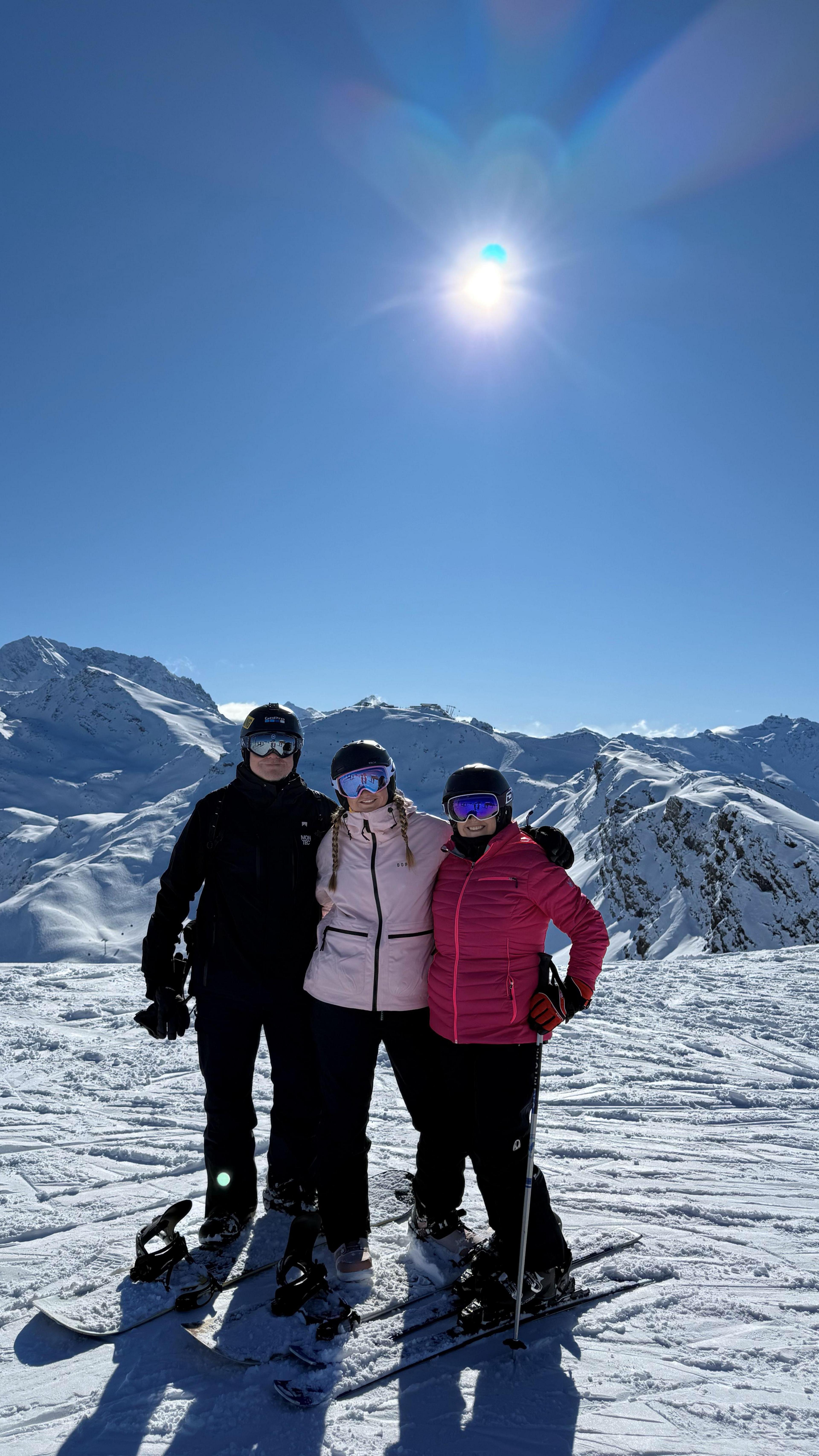 The image shows three people standing together on a snowy mountain landscape under a clear, bright blue sky. All three individuals are wearing winter sports gear, including helmets, goggles with reflective lenses, insulated jackets, and snow trousers, and each is standing on skis or a snowboard. The person on the left stands on a snowboard, while the two individuals on the right are positioned on skis, with one of them holding a ski pole. Behind the group, there are snow-covered mountain peaks and ridges stretching across the background, with the sunlight shining strongly from above and slightly behind them. The bright sun creates sharp shadows on the snow and highlights the crisp texture of the slopes, giving the scene a vivid, high-contrast appearance with clear visibility and expansive scenery.