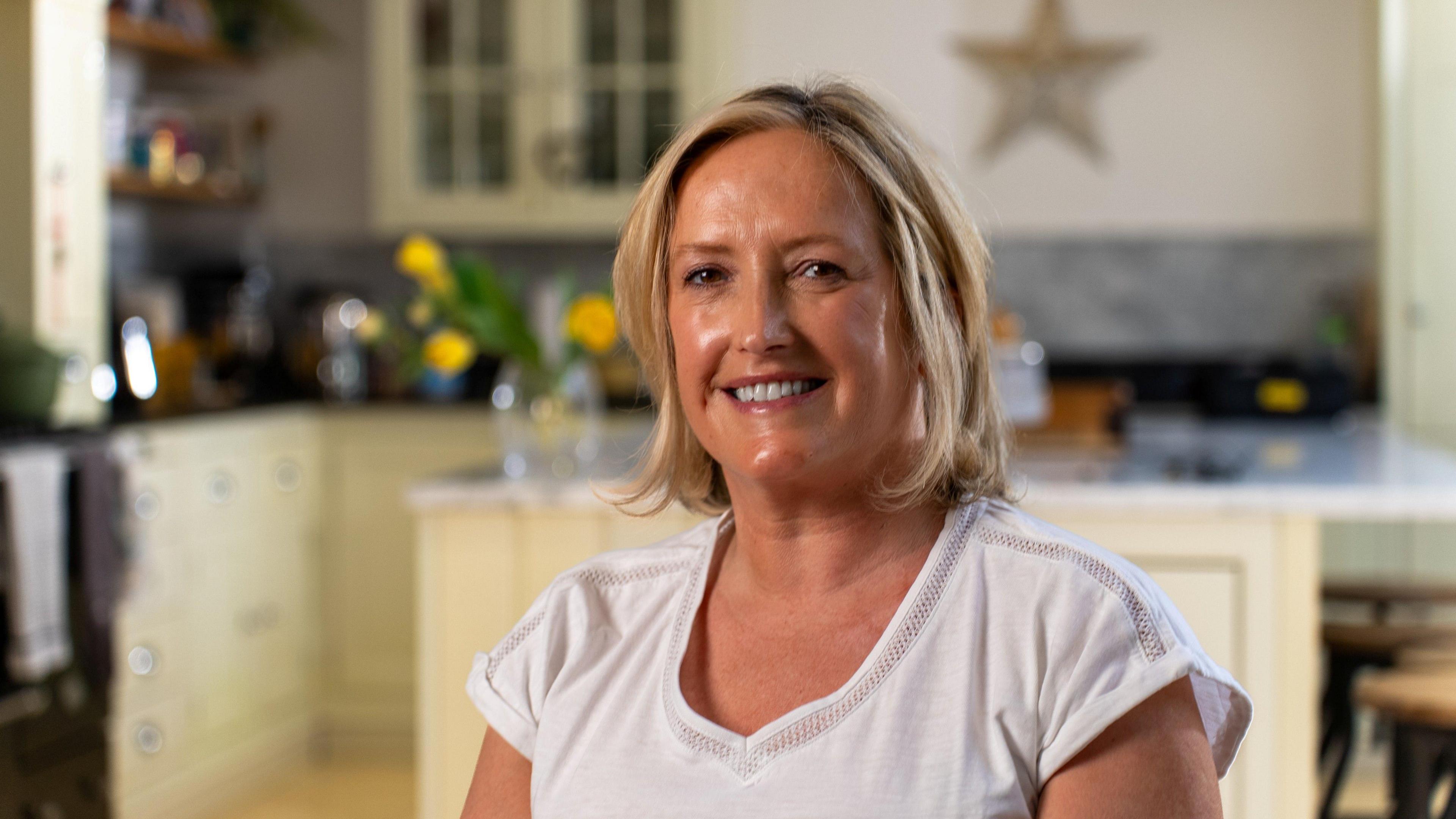 A woman sitting indoors in what appears to be a kitchen setting. She has short, light-coloured hair and is wearing a plain white short-sleeved top, positioned in the foreground and facing the camera. Behind the person, there are light-coloured cabinets, a countertop, and kitchen items such as jars, a kettle, and a vase containing yellow flowers, all slightly out of focus. The lighting is warm and soft, illuminating the person’s face while creating a gentle blur in the background.