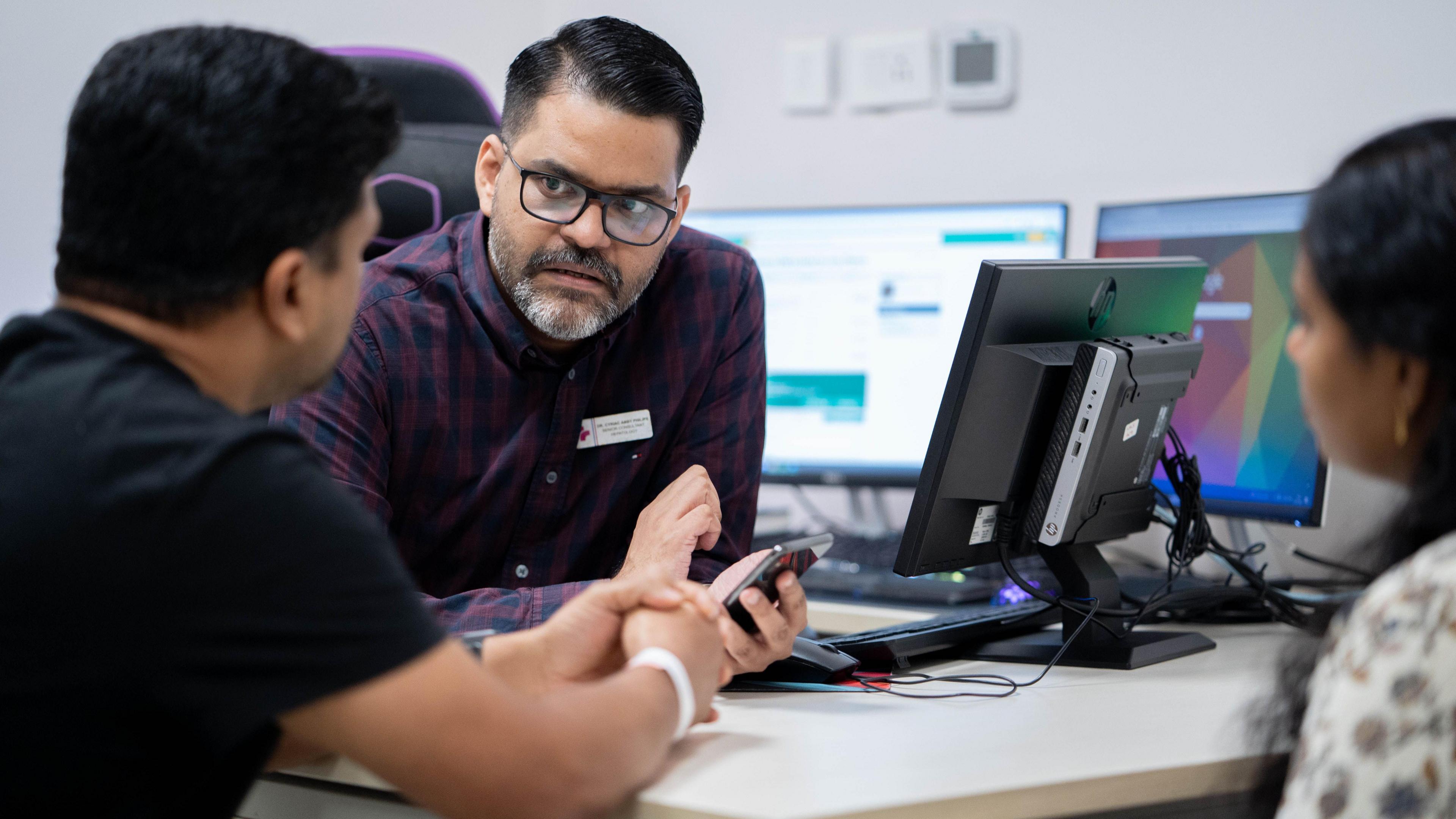 Dr Cyriac Abby Philips, wearing a checkered plum and navy shirt, explains something to his patients on the computer screen 