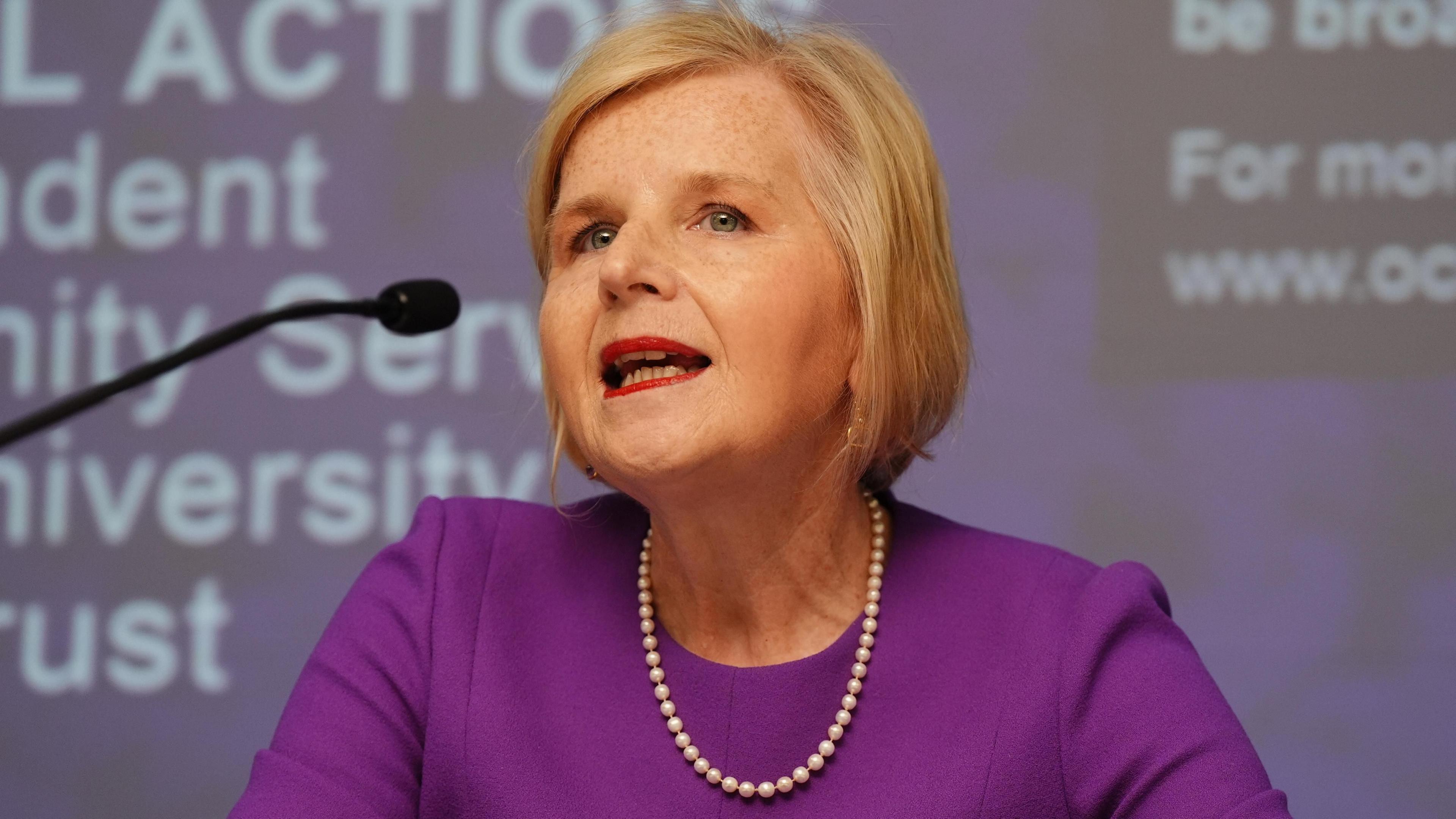 Donna Ockenden speaks into a microphone. She has blonde hair in a bob, is dressed in purple and is wearing pearls