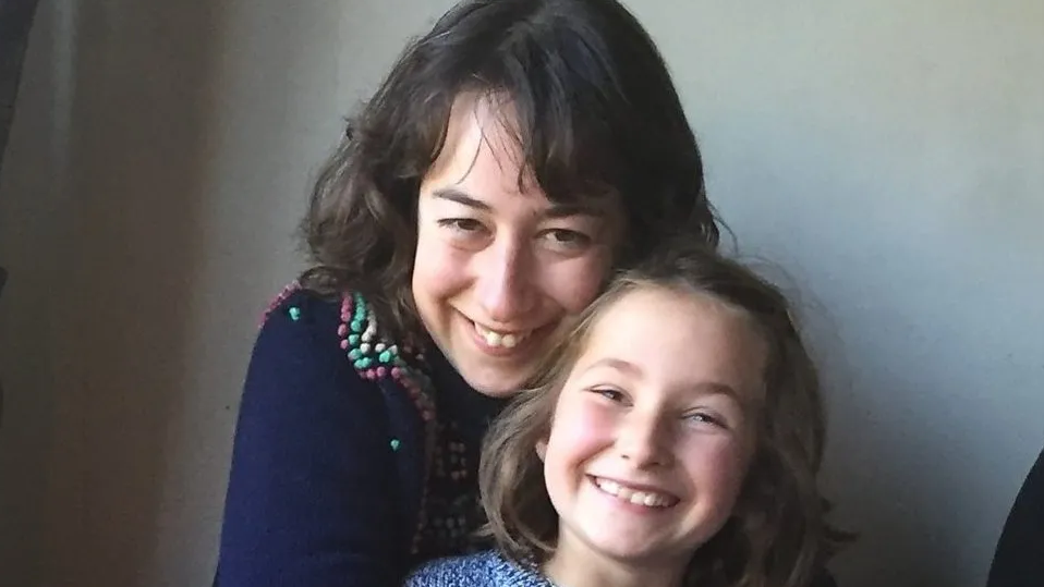 Photo of Martha Mills as a young girl with her mum, Merope, behind her. Merope has long brown hair, wears a dark grey top and is smiling at the camera. Martha has light brown/blonde hair and smiles broadly at the camera. She wears a light blue patterned outfit. 