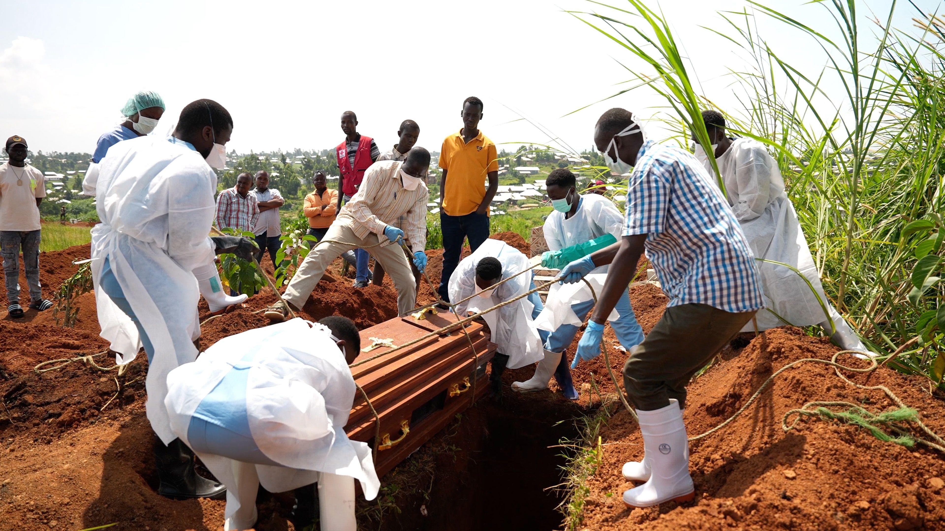 People wearing protective gear, such as masks and gowns, lower a coffin into the ground using ropes