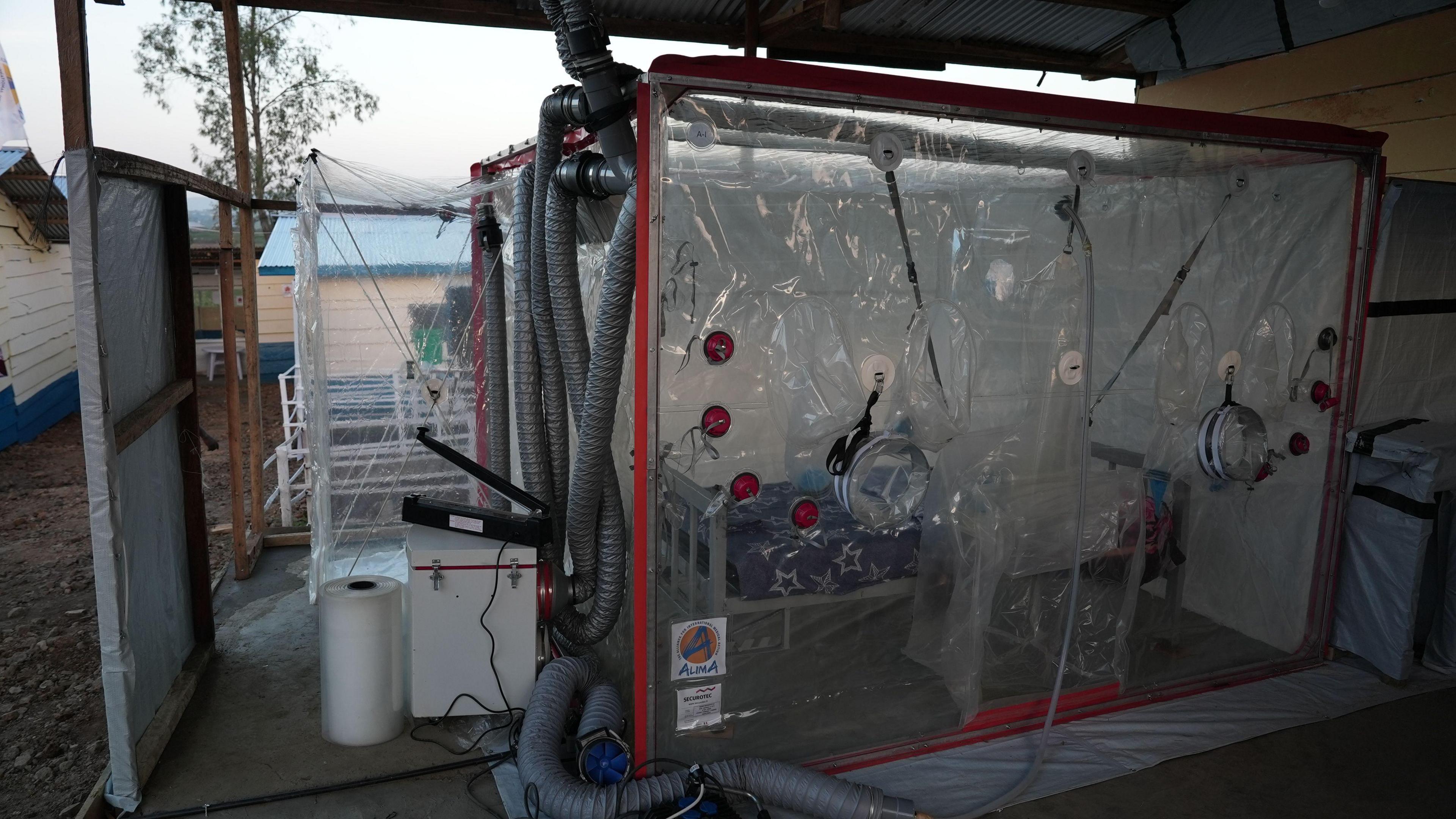 A bed sits inside a plastic cubicle. Pipes protude from the cubicle.