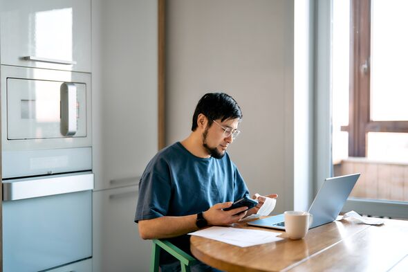 Man in glasses budgeting expenses, using a smartphone and laptop at a wooden table. Managing personal finance and taxes