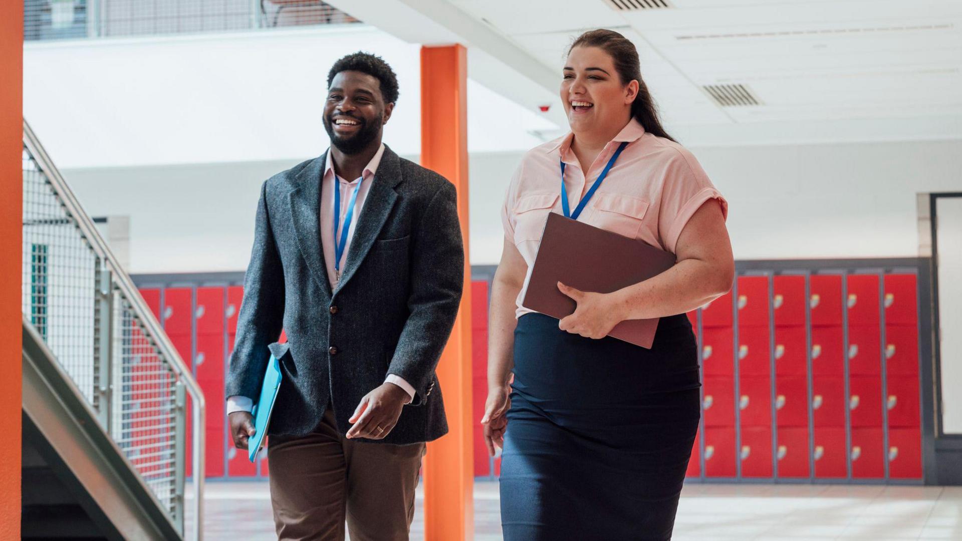Two employees walk together while smiling. They're both wearing lanyards and holding folders.