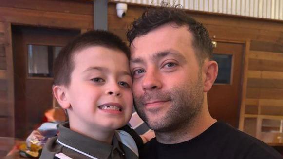 A man and a young boy are cheek to cheek as they smile at the camera. The man has short dark brown hair and is wearing a black T-shirt. The boy also has short dark brown hair and is wearing a dark grey shirt. 
