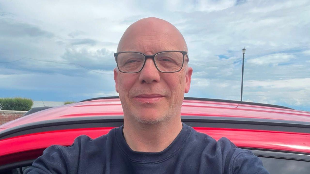 A selfie of a man wearing a navy jumper and glasses. He is standing outside, seemingly in front of a red car. 